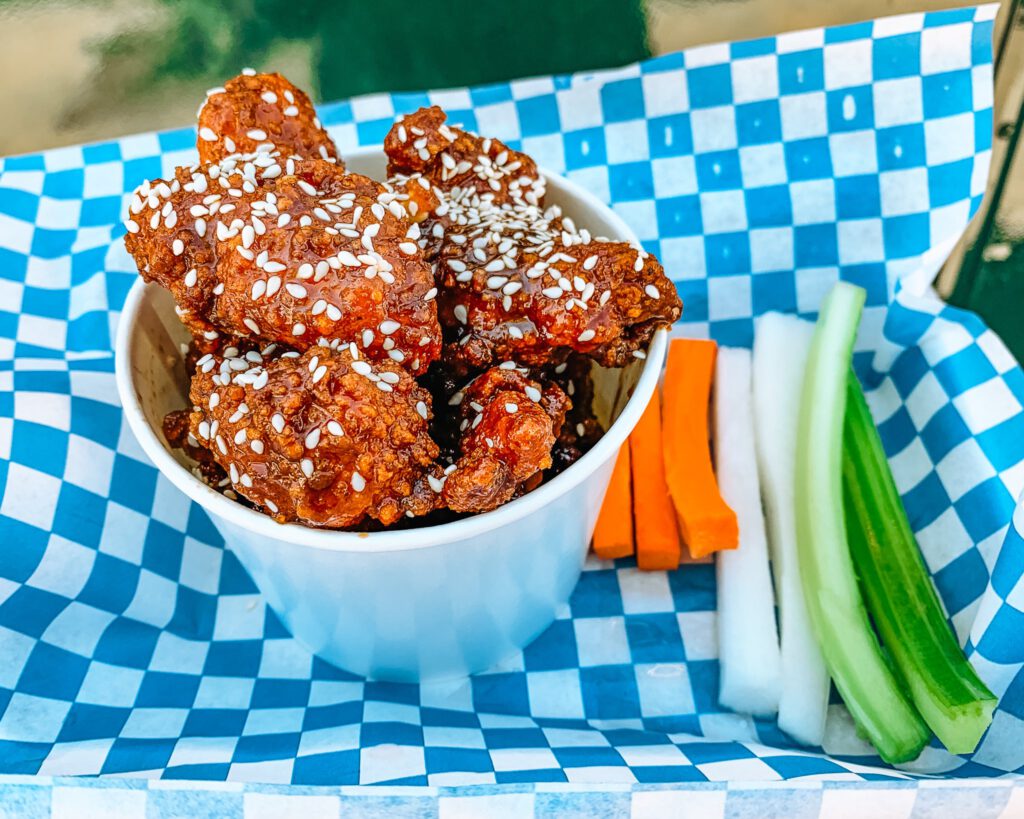 Cup of fried chicken bites tossed in a spicy sauce and topped with sesame seeds sit in a blue and white checkered basket