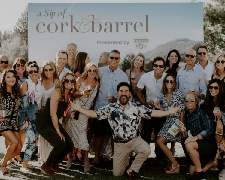 group of 15 men and women stand in front of a step and repeat with the logo cork & barrel while they hold glasses of wine and smiling