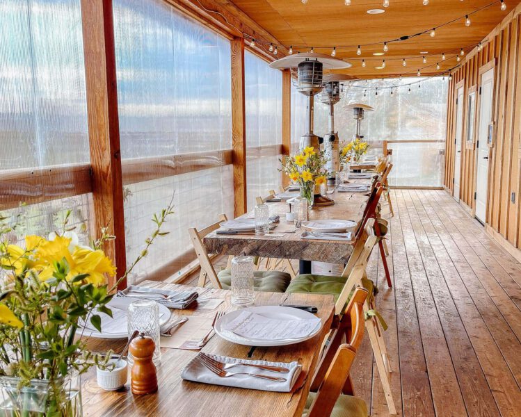 Farm deck is lined with communal wood tables and set with vases of yellow flowers and white plates
