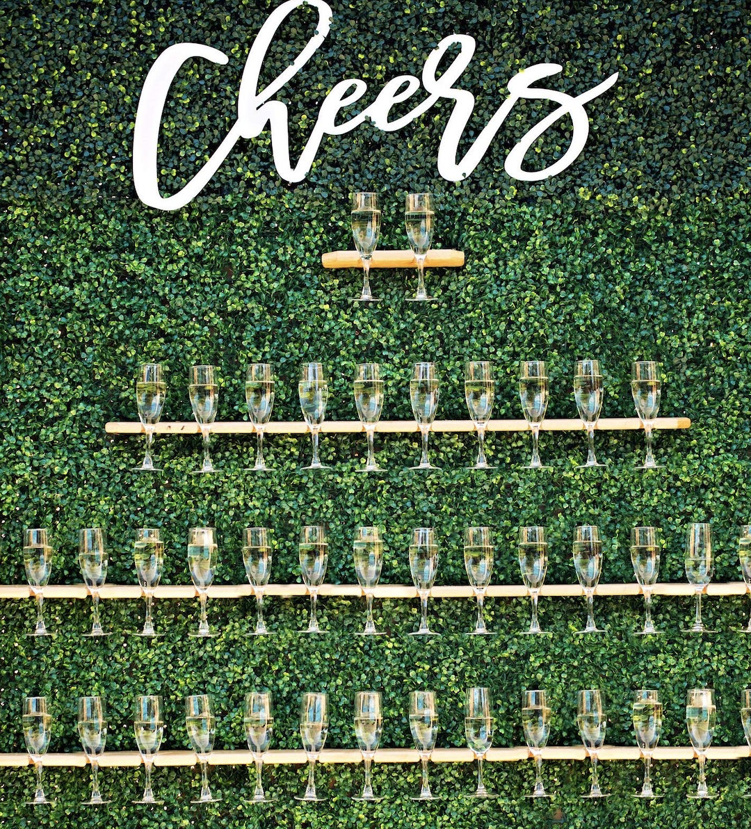 Green wall with 3 rows of shelves holding champagne glasses with the words 'cheers' on a vintage mobile cart