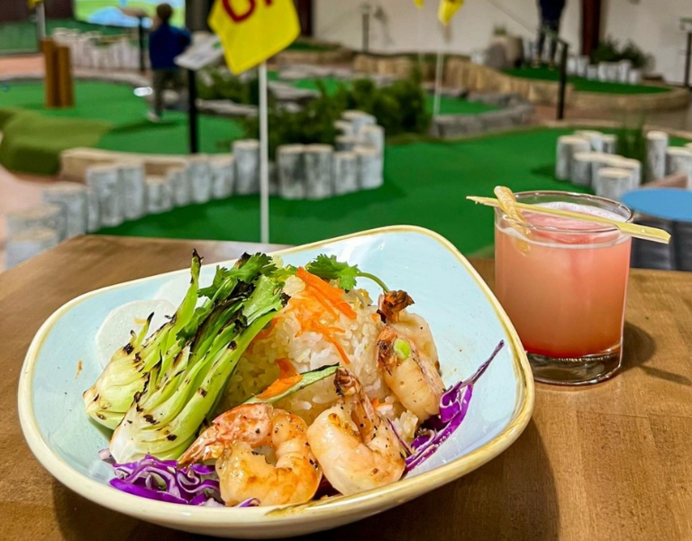 plate of grilled shrimp and a pink cocktail in the foreground with mini golf course at walt reilly's in the background