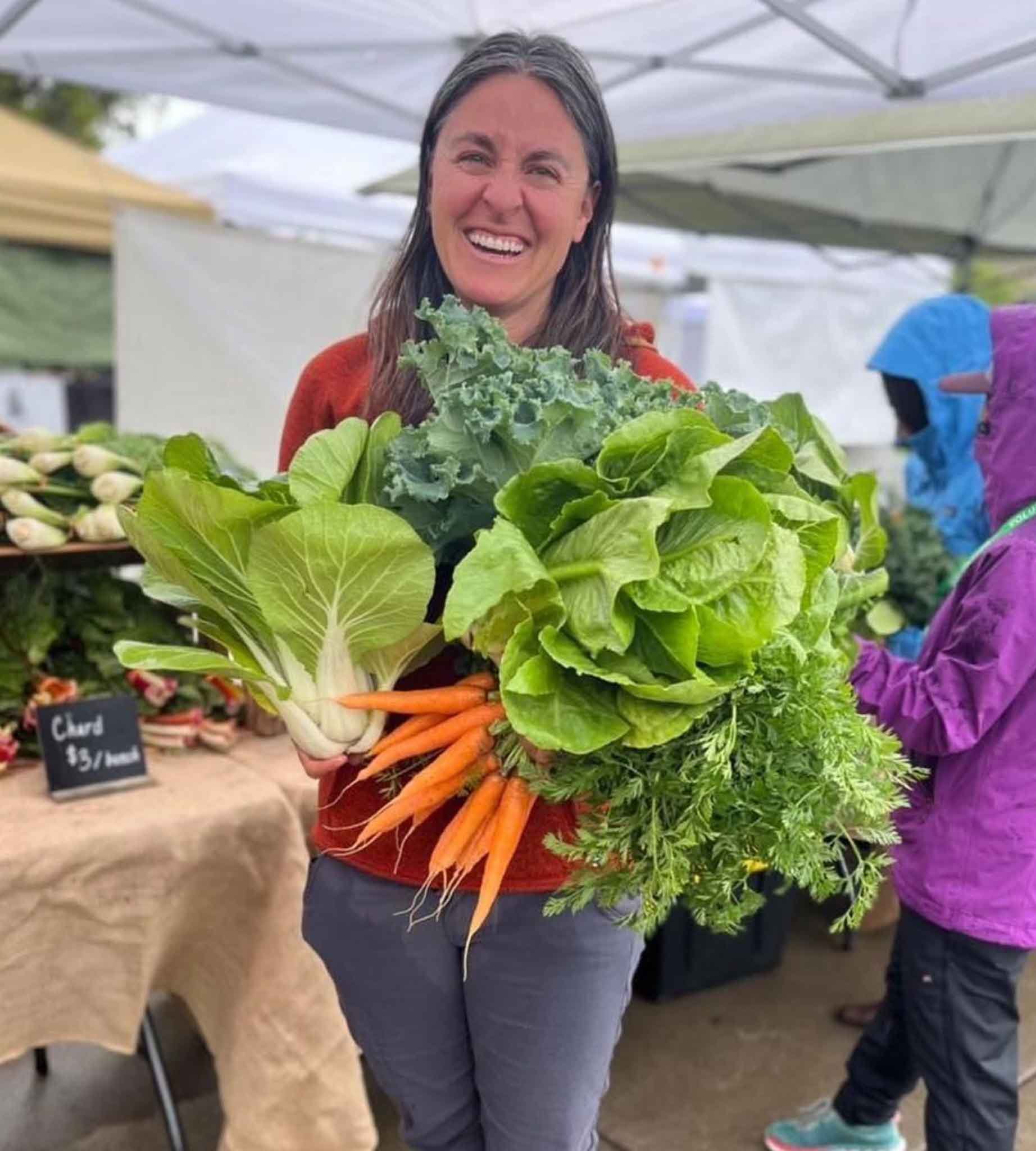 Woman holds a bunch of carrots, head of lettuce, and bok choy at the farmers market in sisters oregon