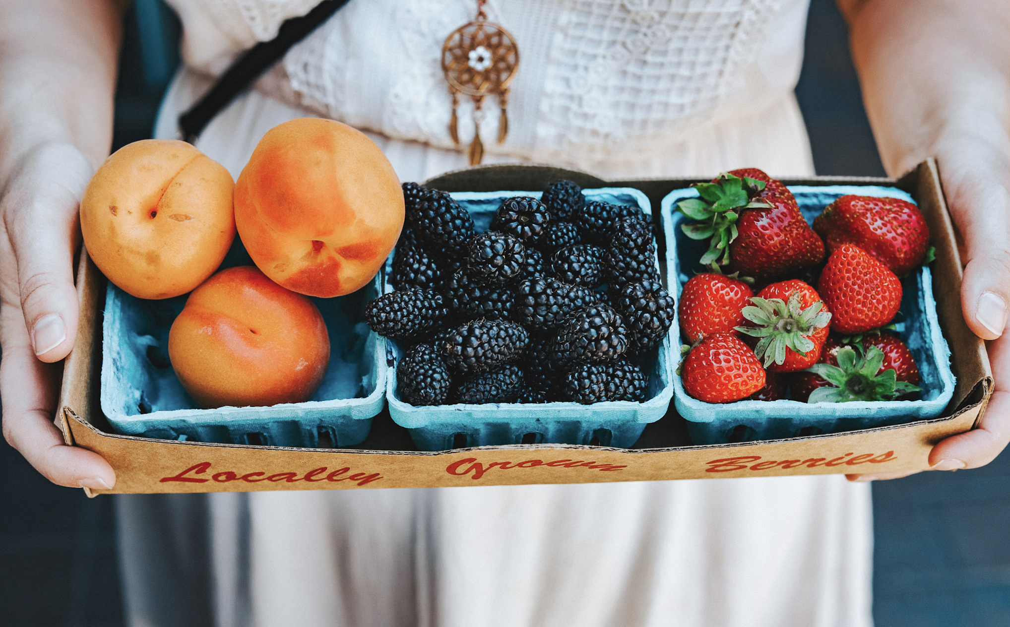 woman's hands hold a box with three trays of apricots, blackberries, and strawberries at a bend oregon farmers market