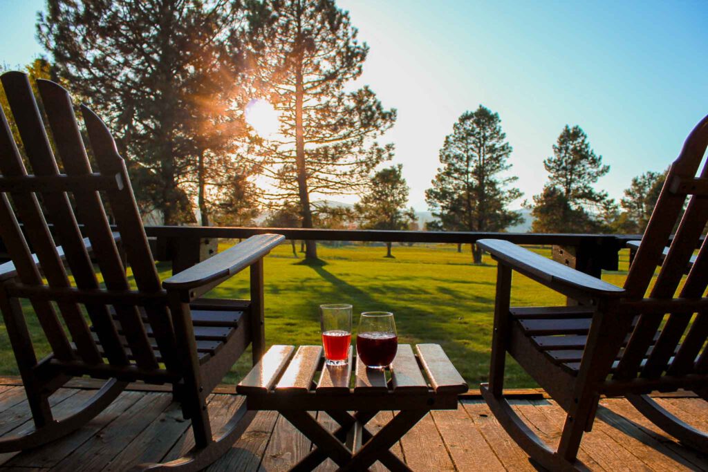 Deck of sunriver resort with two rocking chairs and table with cocktails overlooks green grass with sun setting behind pine trees