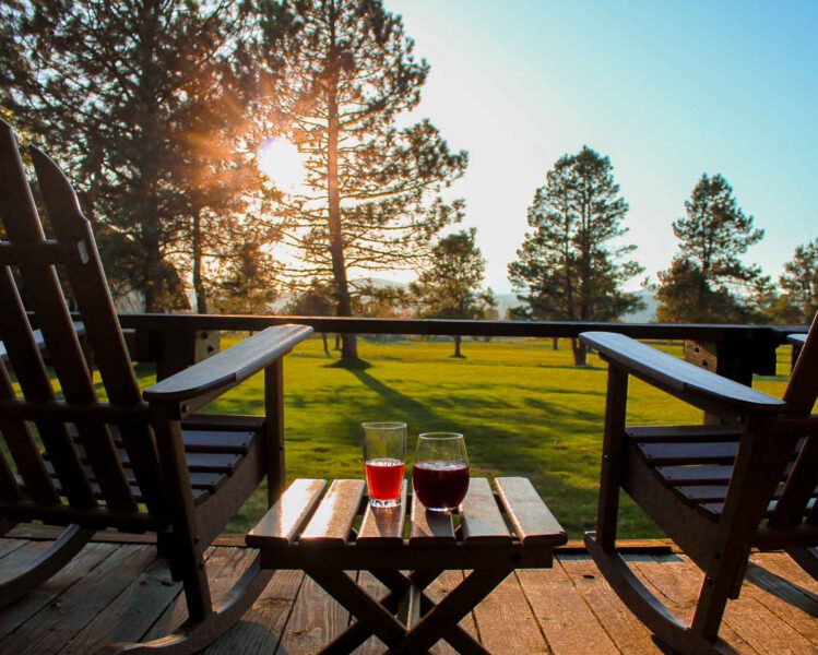 Deck of sunriver resort with two rocking chairs and table with cocktails overlooks green grass with sun setting behind pine trees
