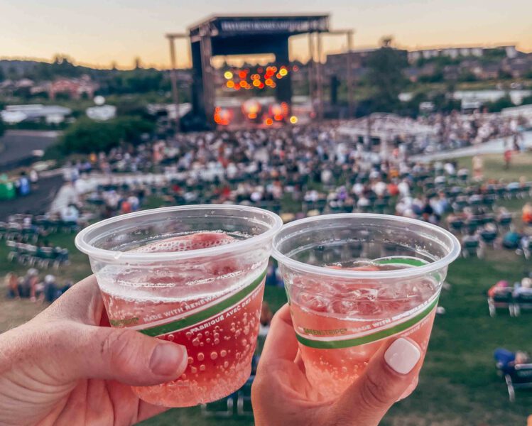 two hands holding cups of sparkling rosé overlooking bend concerts at hayden homes amphitheater