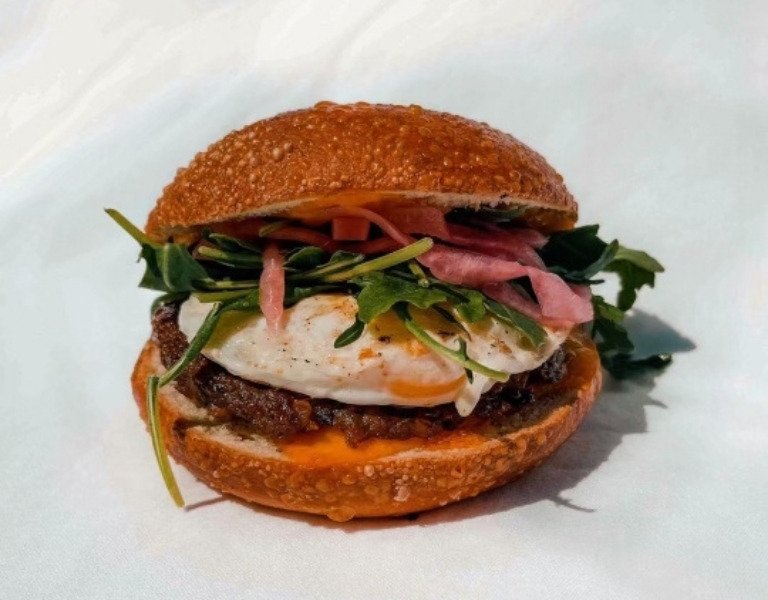 Toasted bagel shows inside sausage patty, soft fried egg, arugula, and pickled onions with a white backdrop at Bo's Falafel Bar in Bend Oregon