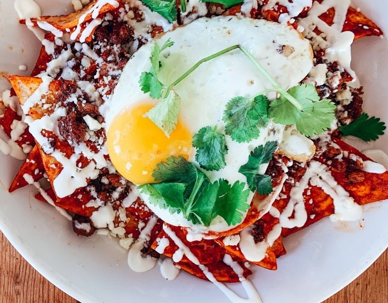 overhead view of a fried egg and cilantro sprigs over red chilaquiles drizzled in sour cream