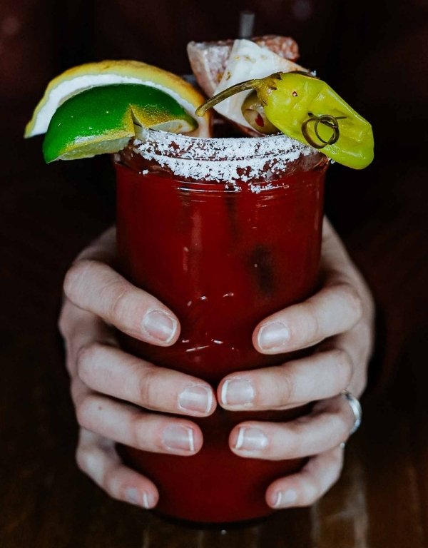 Hands wrapped around a tall mason jar with bloody mary, salted rim, and skewers of lime, lemon, salami, cheese, and pickles