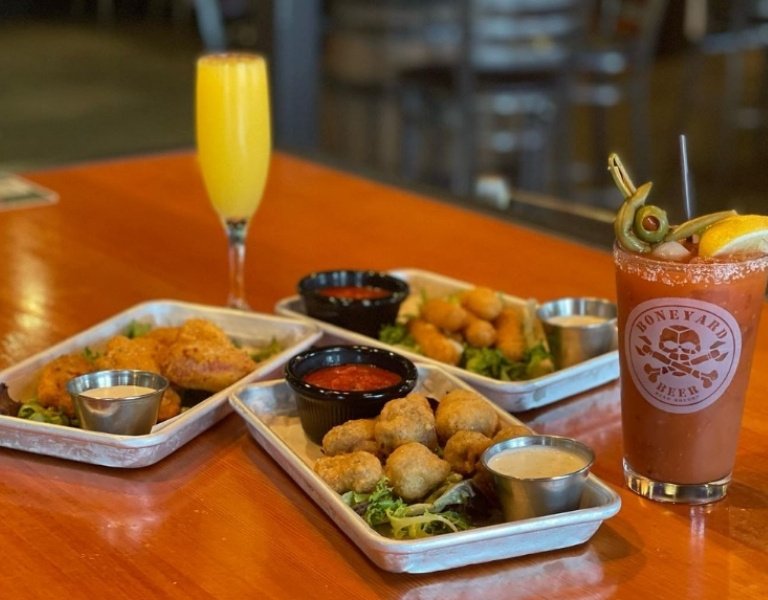 Pub table holds three silver trays of fried sticks and balls, with a bloody mary and mimosa in the background