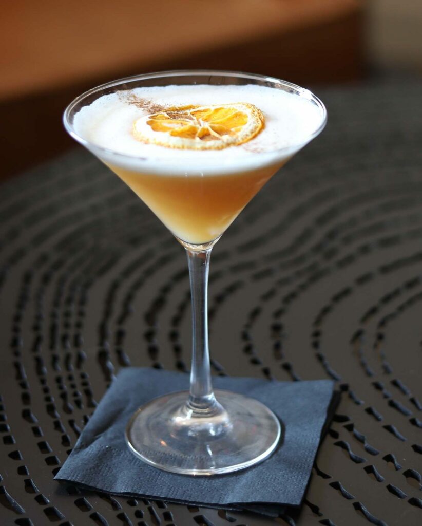 Martini glass contains an orange cocktail with a foamy white top and garnished with a dried orange slice