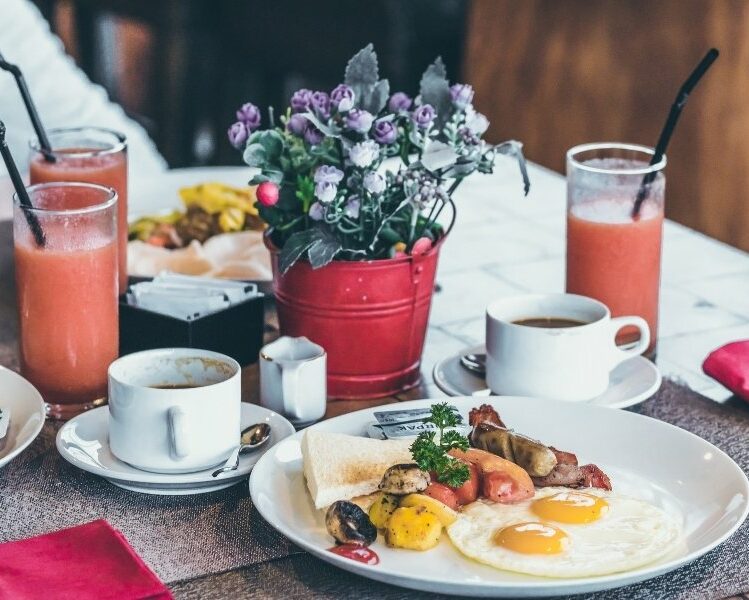 plate of brunch sits next to three glasses of pink juice with a red buket filled with purple flowers in the background
