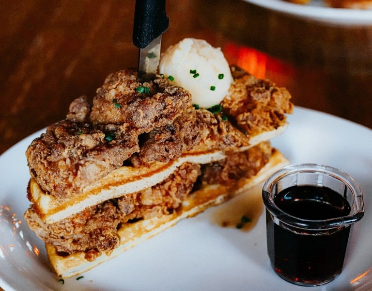 steak knife holds together a stack of belgian waffles and fried chicken, topped with a ball of butter which is listed as best brunch in bend oregon during football season