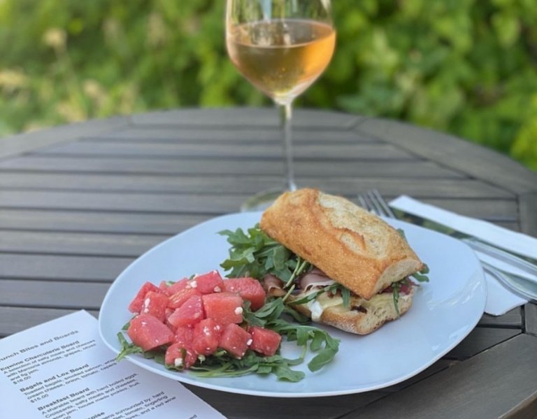 on a white plate sitting on a picnic table: sandwich with arugula and a side of diced watermelon, with a glass of rosé in the background