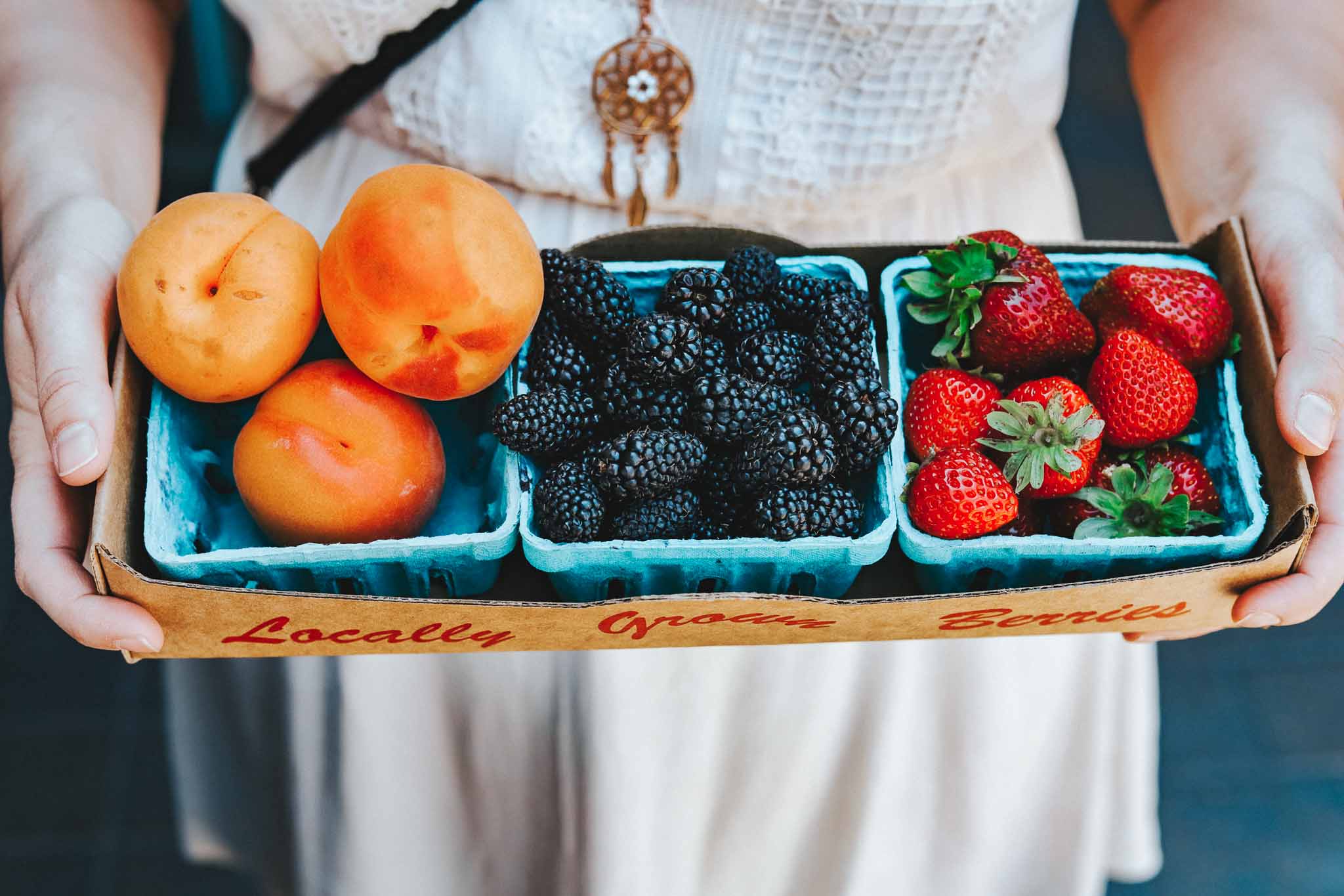 woman's hands hold a box with three trays of apricots, blackberries, and strawberries at a bend oregon farmers market