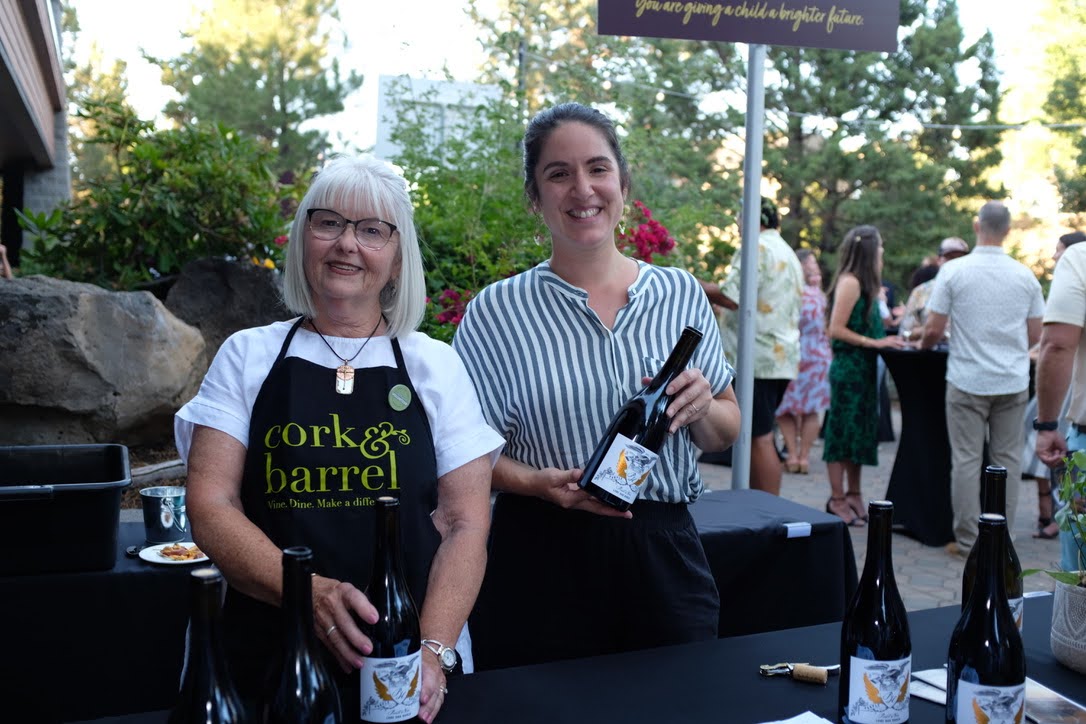 two women hold a bottle of wine wearing cork & barrel aprons while volunteering