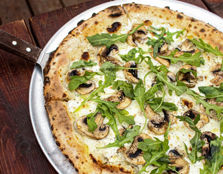 Pizza with mushrooms and arugula sits in a pan with a pizza slicer wedged beneath a slice