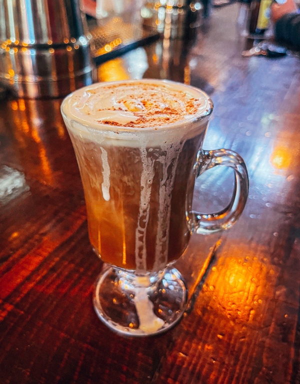 Irish coffee in a clear glass mug sits on the bar at River Pig Saloon.