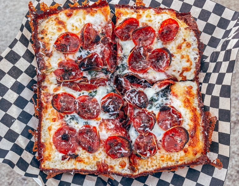 Square Sicilian style pizza is topped with pepperoni and placed in a basket lined with black and white checkered paper