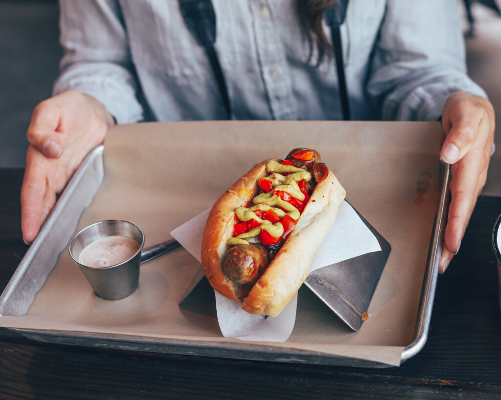 sausage in a bun covered with red peppers and yellow mustard sits on a tray held by womans hands.