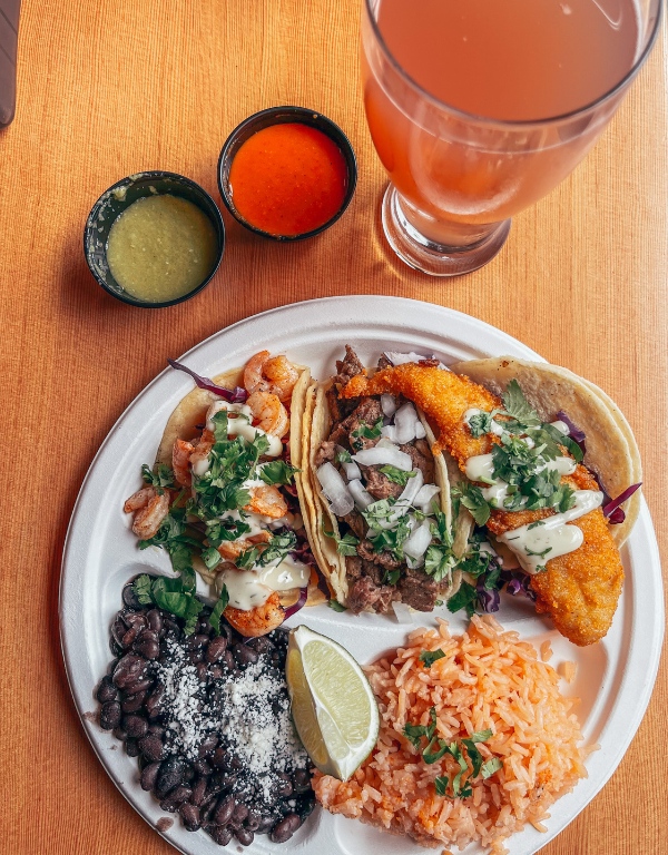 overhead view of plate with three tacos, black beans, rice, and pint of kombucha off to the right.