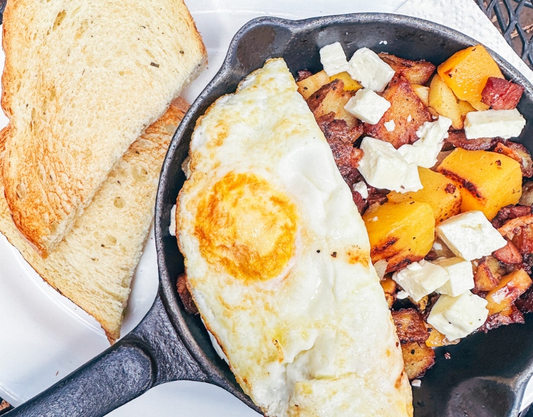 small cast iron skillet with roasted butternut squash and apples topped with a fried egg next to a plate of sliced sourdough toast